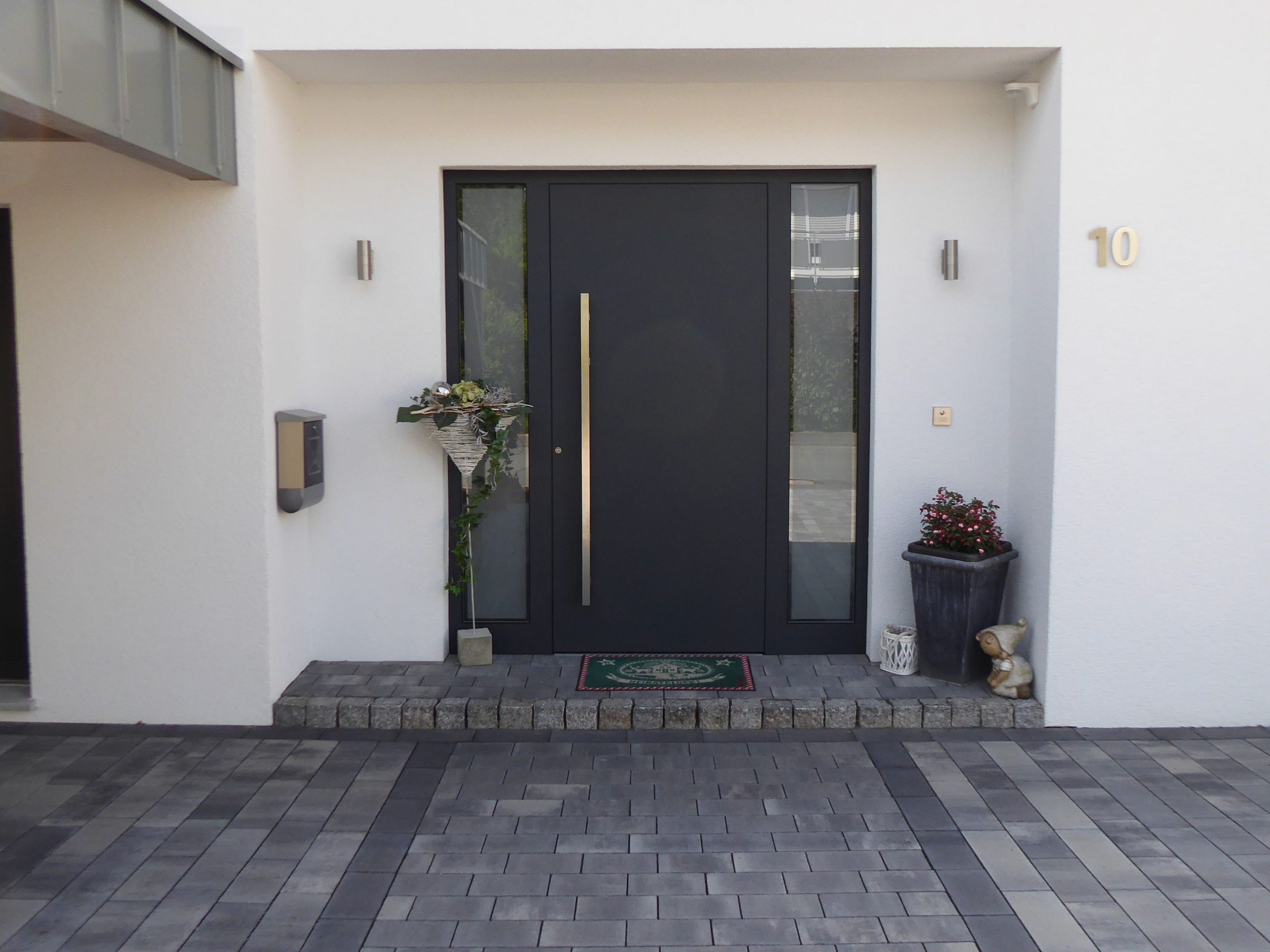 Detailansicht des Hauseingangs eines Einfamilienhauses mit dunkler Haustür, seitlichem Glasfeld und gepflastertem Vorbereich