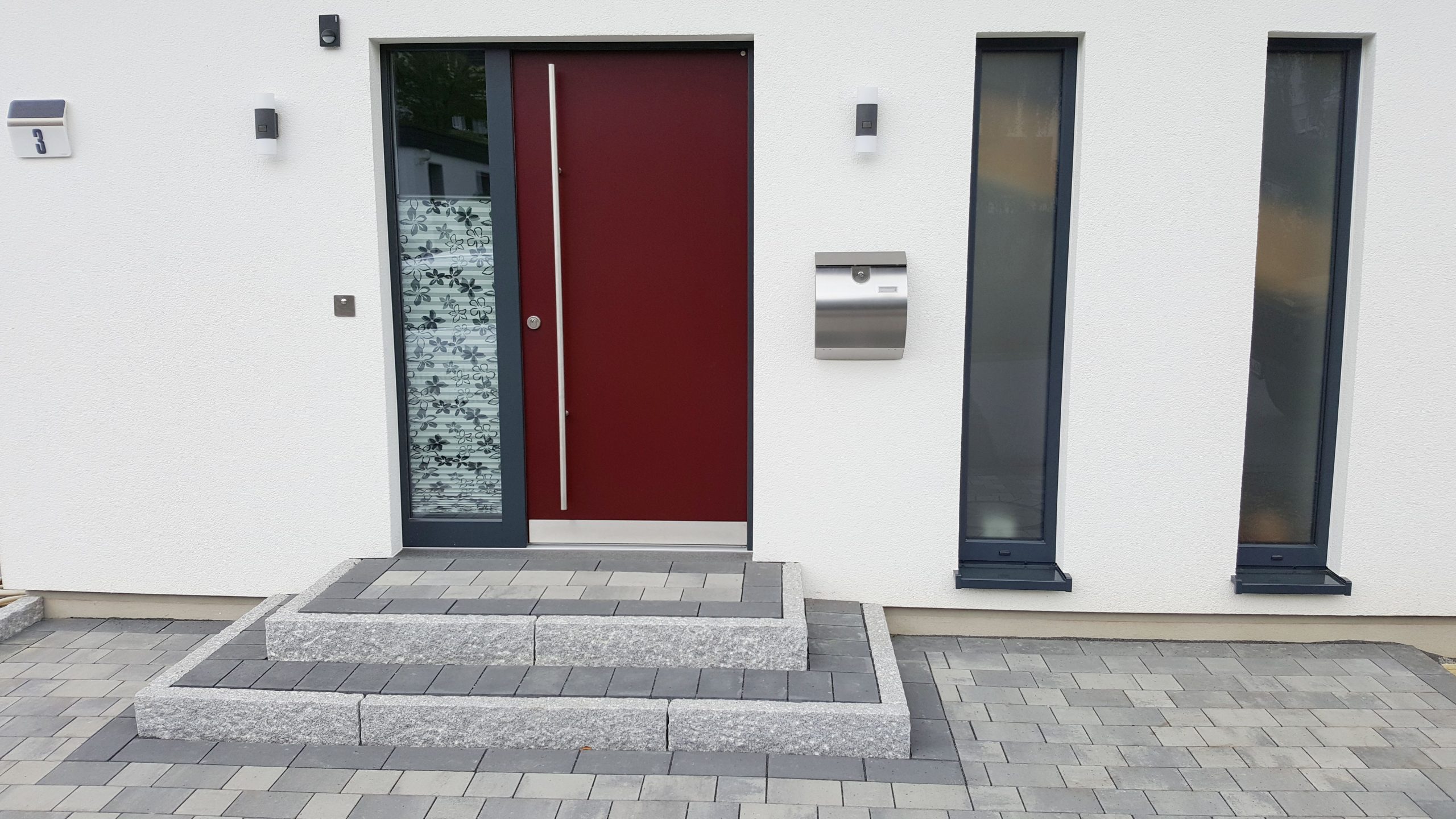 Hauseingang mit dunkelroter Tür, seitlichem Glasfeld und heller Putzfassade