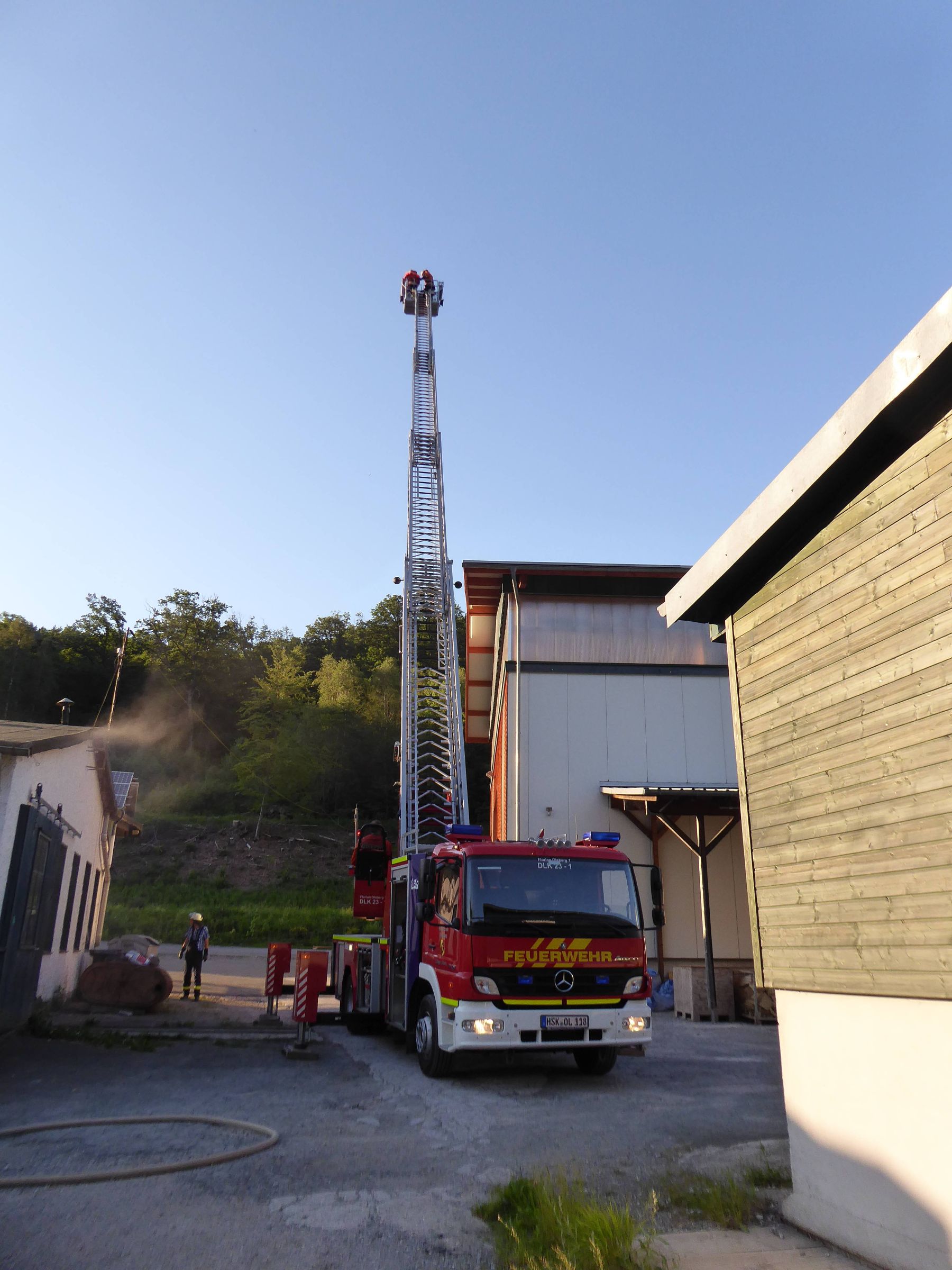 Feuerwehrfahrzeug mit ausgefahrener Drehleiter steht auf einem Betriebsgelände zwischen Hallengebäuden