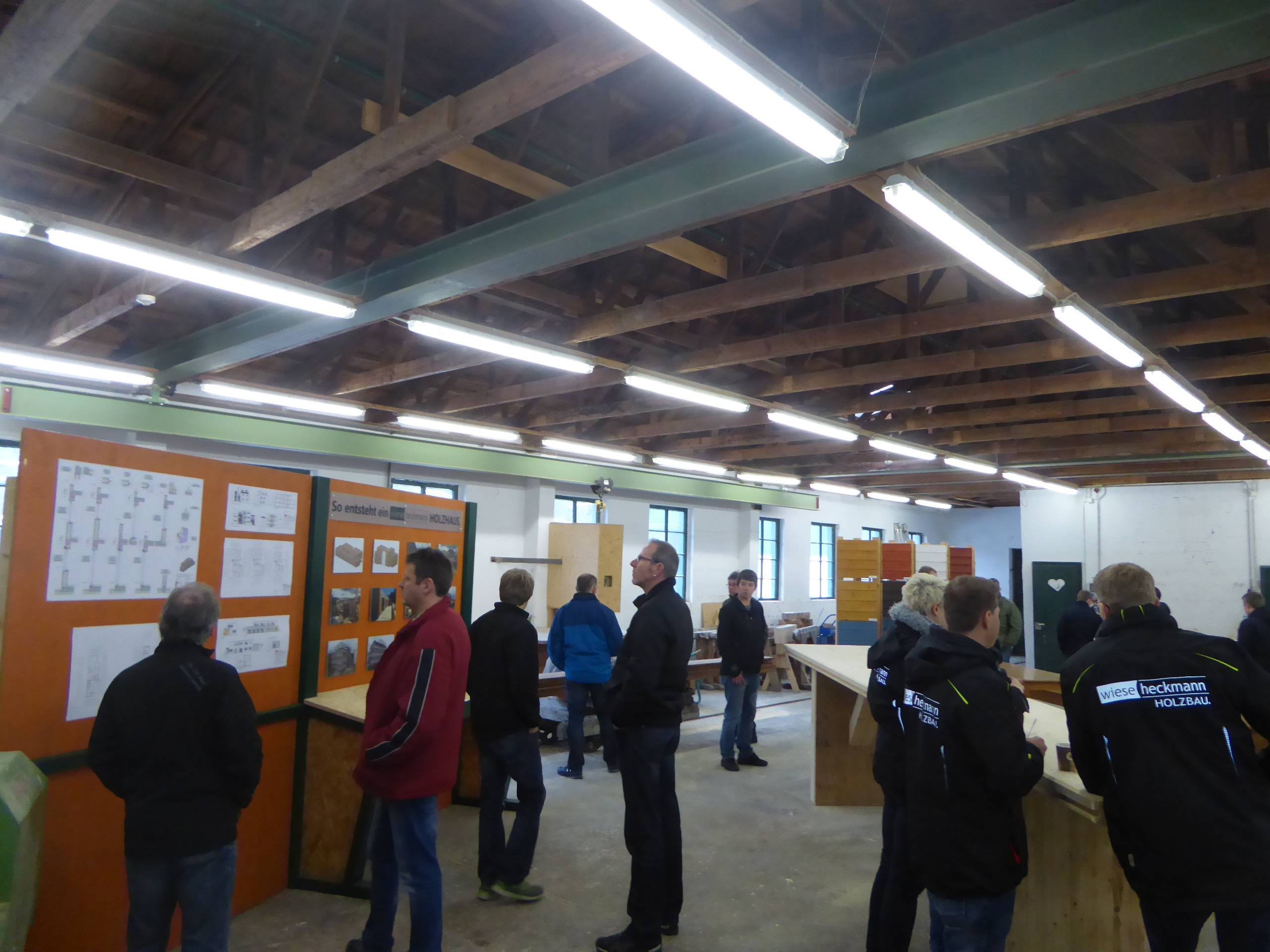 Besucher stehen in einer Werkhalle und betrachten Pläne sowie Holzbauelemente